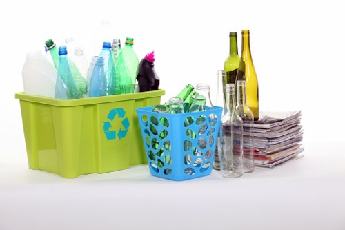 A collection of various recyclable glass bottles and plastic containers placed in a green plastic bin with a recycling symbol, along with a smaller blue basket containing additional glass bottles, set against a plain white background. To the right, there are more glass bottles of different shapes and colors, including clear, green, and amber, some with long necks, stacked next to a neat pile of newspapers or magazines. The scene appears to be a depiction of waste or rubbish ready for disposal or collection, emphasizing recycling materials commonly handled during rubbish removal services. The environment suggests an indoor setting, possibly a workspace or designated rubbish collection area, and the items are arranged in an orderly fashion, highlighting proper separation of recyclable waste. This setup aligns with services provided by Garden Clearance Plaistow, focusing on waste management and rubbish removal, particularly involving glass and paper recyclables, within the Plaistow postcode area.