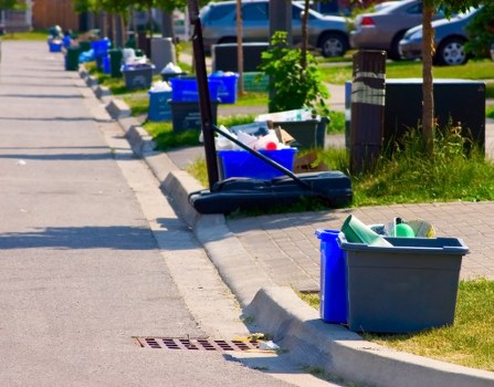A black push broom with a long handle and bristles resting on the pavement next to a grey plastic bin filled with green and blue objects, situated along a residential sidewalk. Several blue bins are lined up along the edge of the pavement, containing various discarded items, and the background features parked cars, small trees, and a grassy verge, indicating a suburban area. The scene appears to be part of a rubbish removal or street cleaning effort in a neighbourhood potentially serviced by Garden Clearance Plaistow, with a focus on waste collection and tidiness along the street. The environment is well-lit, with natural daylight casting shadows on the ground, and the overall setting reflects typical urban or suburban rubbish disposal activities in the UK.