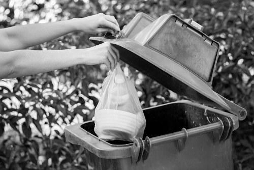 A person’s hands are seen lifting the lid of a large, dark grey outdoor waste bin situated on a paved driveway or garden area. The bin’s lid is open to reveal a partially filled clear plastic bag containing white household items or waste. The bin has a sturdy, textured surface with visible hinges on the side. Surrounding the bin are leafy bushes or hedges, indicating a garden setting common in residential areas such as Plaistow. The scene is captured in natural light, emphasizing the functional purpose of rubbish collection and disposal services provided by Garden Clearance Plaistow. The image highlights the process of rubbish removal, with the bin positioned in an accessible outdoor space suitable for garden or household waste clearance.