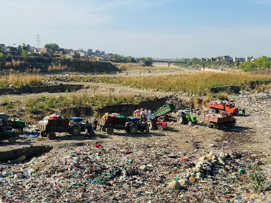 A small vintage blue flatbed truck parked parallel to a pavement, loaded with a variety of waste materials including cardboard boxes, black plastic bags, and miscellaneous debris, secured with ropes a