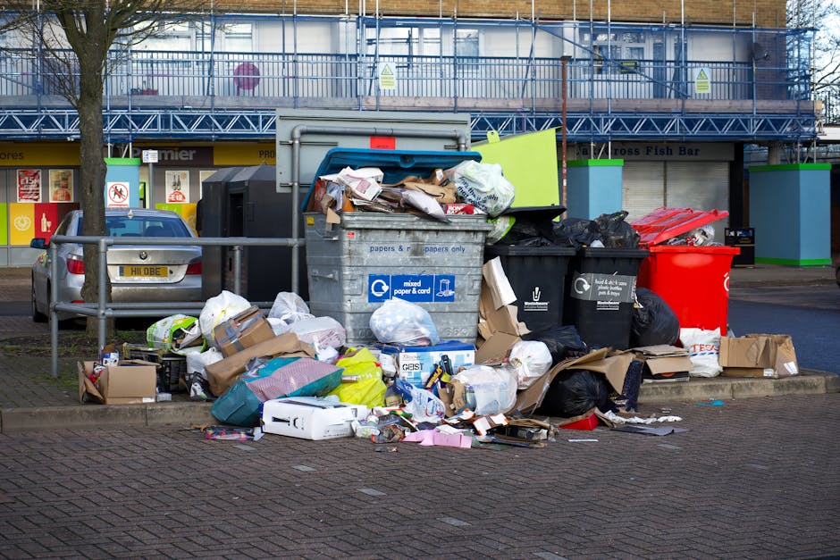 A collection of overflowing rubbish bins and waste bags piled on a paved sidewalk in front of a commercial retail area. The collection includes a large metal mixed paper and cardboard bin with its lid
